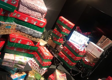 Ein Raum voller weihnachtlich verpackter Geschenkkartons, viele in rot-grünen Farben, einige mit Mustern und Schleifen. Die Päckchen sind aufgestapelt auf Stühlen, Tischen und dem Boden. In der Mitte steht ein Leuchtkasten mit der Aufschrift „XMAS IN A BOX – FRANKFURT“. Im Hintergrund moderne Wanddekoration mit Bildern („Born to be wild“, Ananas) und eine Deckenlampe mit drei Glühbirnen.