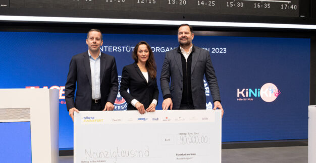 Drei Personen stehen nebeneinander und halten einen großen Scheck über 90.000 Euro. Der Hintergrund zeigt eine blaue Wand mit verschiedenen Logos und einem digitalen Zeitplan. Der Scheck ist von der Börse Frankfurt und verschiedenen Partnern und wurde für eine Unterstützungsorganisation überreicht.