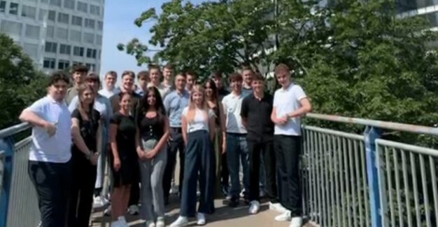 Herzlich willkommen im MRH Trowe Team – Ausbildungsstart 2024! Eine Gruppe von Menschen steht auf einer Brücke und posiert für ein Foto. Im Hintergrund sind grüne Bäume und moderne Gebäude zu sehen. Die Sonne scheint und das Wetter scheint angenehm zu sein.