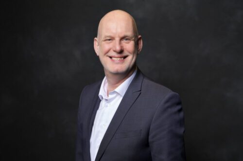 Portrait of Matthias Edelmann, member of the supervisory board, wearing a suit and smiling warmly at the camera, with a dark background.