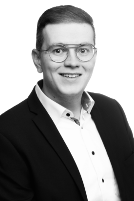 Porträt eines lächelnden jungen Mannes mit Brille im Business-Outfit – dunkles Sakko, weißes Hemd, neutraler Hintergrund (schwarz-weiß Aufnahme).