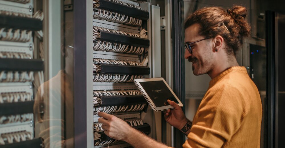 IT-Techniker mit Tablet in der Hand überprüft Kabel und Anschlüsse in einem geöffneten Serverschrank, Symbolbild für Elektronik, Netzwerktechnik und Absicherung von IT-Infrastrukturen.