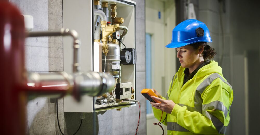 Servicetechnikerin mit Schutzhelm und Warnjacke überprüft Heizungs- oder Industrieanlage mit Messgerät, Symbolbild für Wartung, technische Sicherheit und Maschinenversicherung.