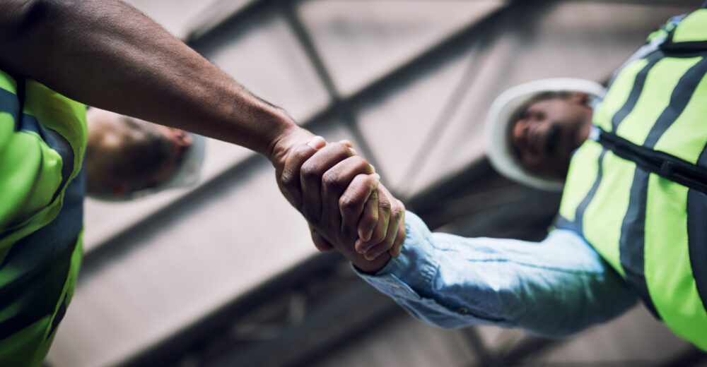 Zwei Bauarbeiter in Schutzwesten und Helmen geben sich die Hand – Symbolbild für Vertrauen und Absicherung durch Montageversicherung.