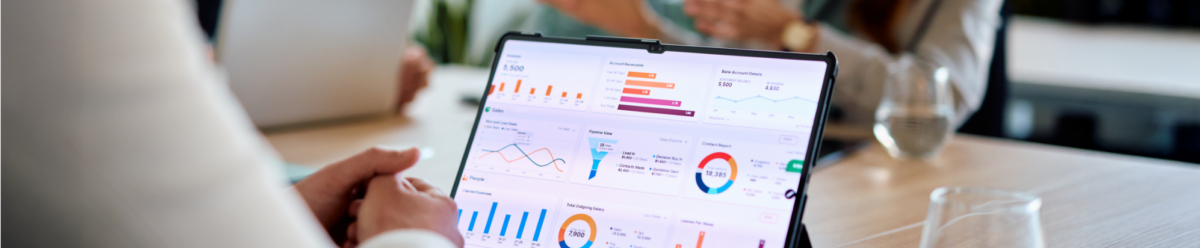 Tablet mit Finanzdiagrammen und Statistiken auf einem Besprechungstisch, im Hintergrund Personen in einem Meeting.