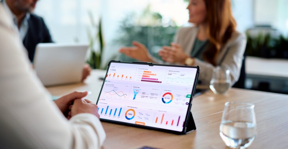 Tablet mit Finanzdiagrammen und Statistiken auf einem Besprechungstisch, im Hintergrund Personen in einem Meeting.