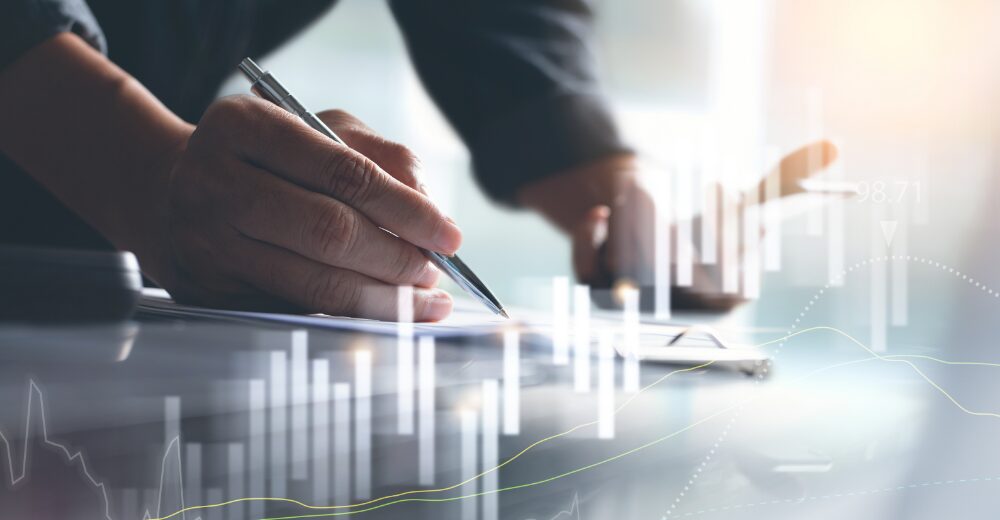 Nahaufnahme einer Hand, die mit einem Stift auf Dokumenten schreibt, überlagert von abstrakten Finanz- und Diagrammgrafiken.
