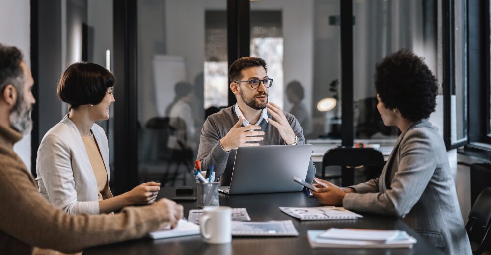 Vier Personen sitzen in einem modernen Besprechungsraum, eine Person erklärt etwas gestikulierend vor einem Laptop.