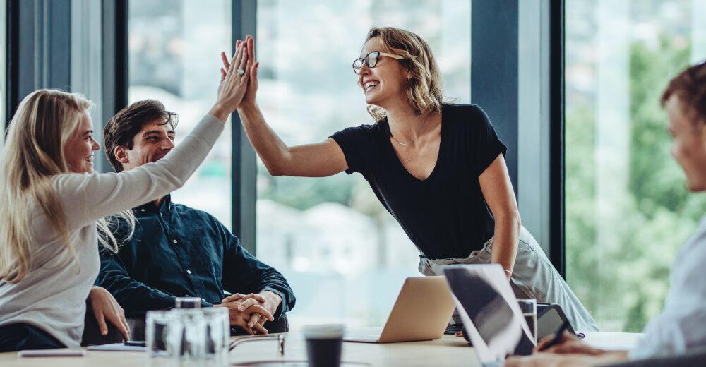 Vier Personen in einem modernen Büro, zwei davon geben sich lächelnd ein High Five in einer Teamsituation.