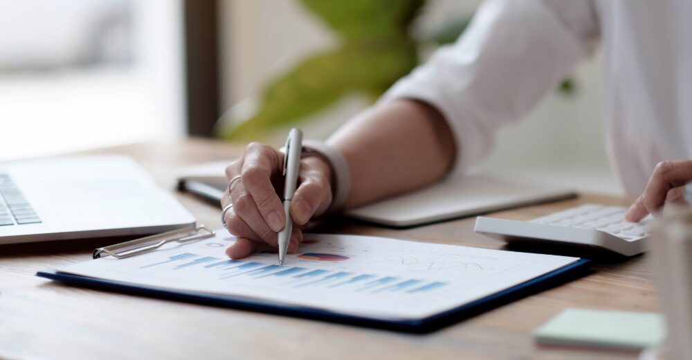 Nahaufnahme einer Hand, die anhand von Diagrammen und Tabellen auf einem Dokument arbeitet, daneben ein Laptop und ein Taschenrechner.