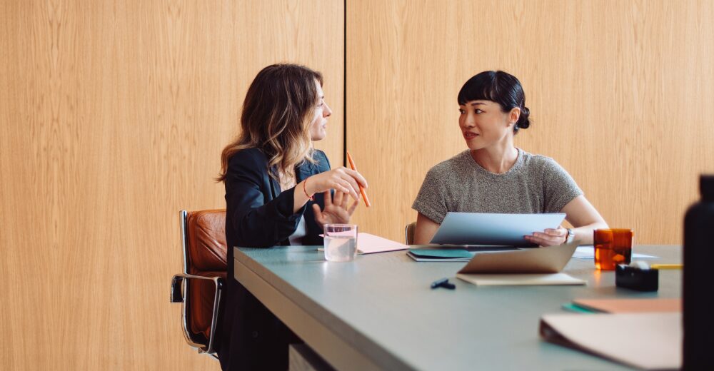 Zwei Personen sitzen in einem modernen Besprechungsraum an einem Tisch und sprechen über Unterlagen.
