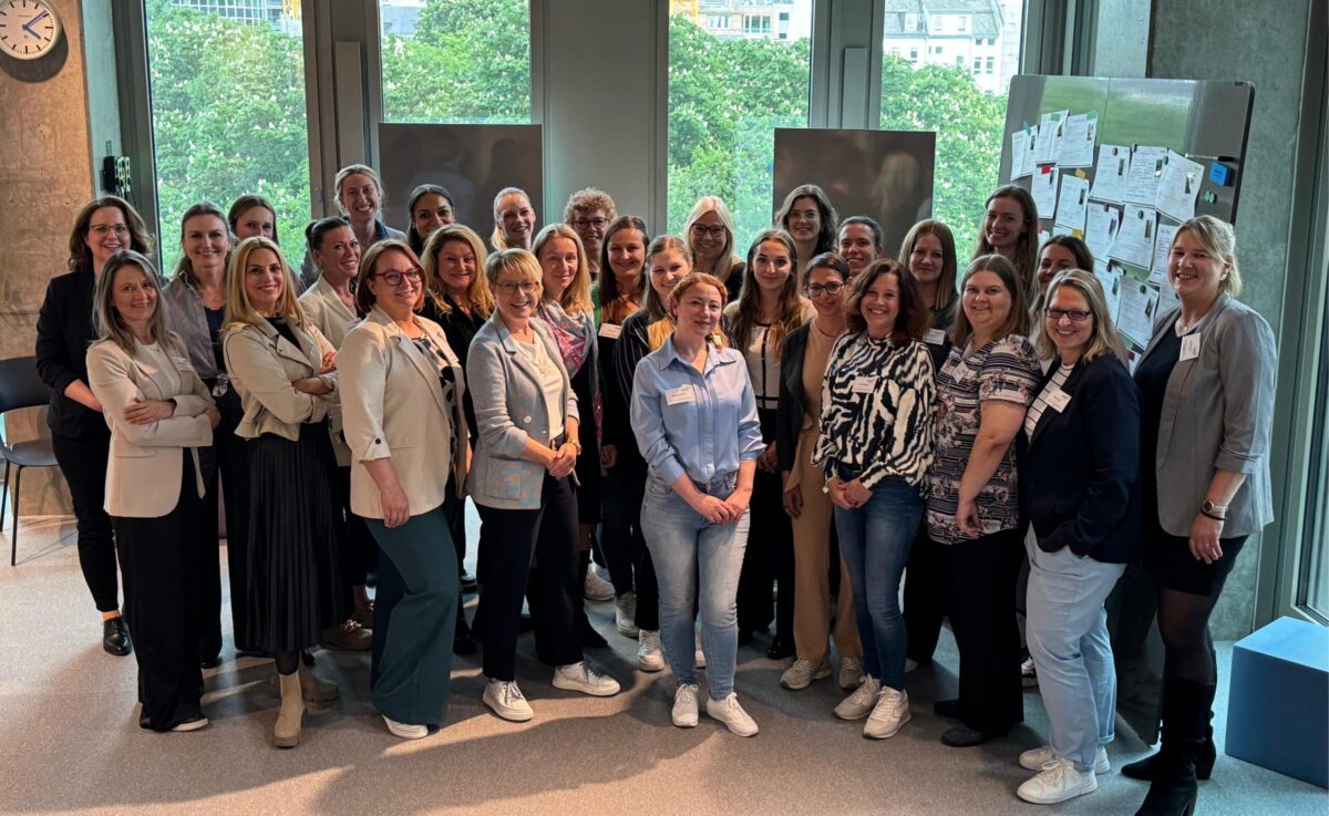 „Gruppenfoto von rund 30 Frauen in einem modernen, hellen Raum vor großen Fenstern. Sie stehen dicht beieinander, lächeln in die Kamera und tragen überwiegend Business-Casual-Outfits. Im Hintergrund sind Bäume sowie einige Pinnwände mit Notizen zu sehen.