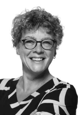 Black-and-white portrait of a smiling woman with short, curly hair and round glasses. She is wearing a patterned top and is looking directly into the camera.