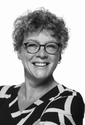 Schwarz-weiß-Porträt einer lächelnden Frau mit kurzen, lockigen Haaren und runder Brille. Sie trägt ein gemustertes Oberteil und blickt direkt in die Kamera.