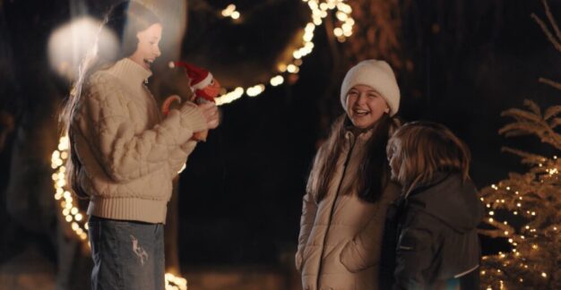 Eine winterliche Szene bei Nacht, in der eine Person einen kleinen Stoffdrachen mit Weihnachtsmütze hält. Im Hintergrund sind Lichterketten und ein dekorierter Weihnachtsbaum zu sehen, die eine warme und festliche Atmosphäre schaffen. Die Personen tragen warme Kleidung, passend zur kalten Jahreszeit.