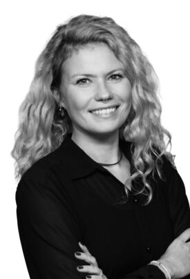 Porträt einer lächelnden Frau mit lockigem, schulterlangem Haar, Nasenring und dunkler Bluse. Sie steht mit verschränkten Armen vor hellem Hintergrund.