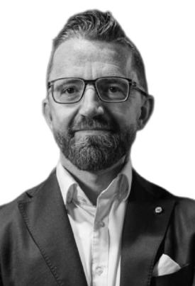 Porträtfoto eines Mannes mit kurzem Haar und Vollbart. Er trägt eine Brille, ein weißes Hemd und ein Sakko mit Einstecktuch. Der Hintergrund ist neutral, und er blickt direkt in die Kamera.