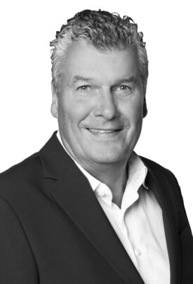 Porträtfoto eines lächelnden Mannes im Anzug und Hemd vor neutralem Hintergrund. Er hat lockiges, kurz geschnittenes Haar und blickt direkt in die Kamera.