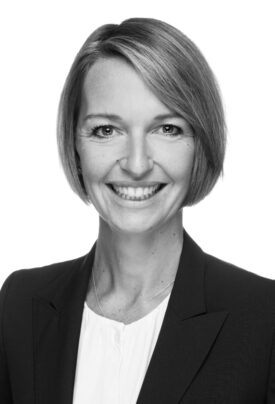 Porträtfoto einer lächelnden Frau mit kurzem, seitlich geteiltem Haar. Sie trägt eine Bluse und ein Sakko und blickt direkt in die Kamera. Der Hintergrund ist neutral.