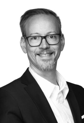 Porträt eines lächelnden Mannes mit Brille und kurzem Bart im schwarzen Anzug vor hellem Hintergrund