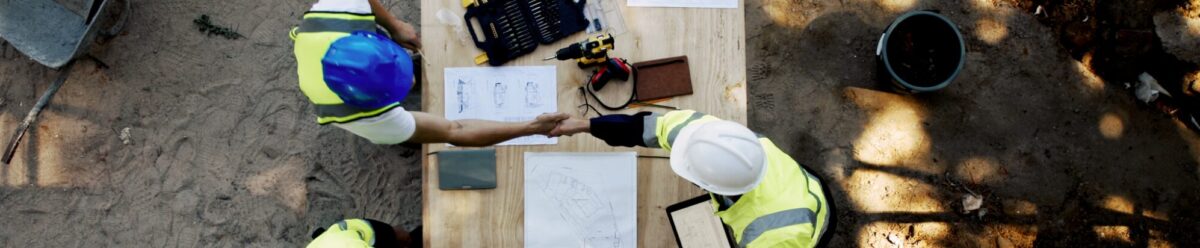 Draufsicht auf drei Bauarbeiter mit Schutzhelmen, die an einem Tisch Baupläne prüfen und sich über ein Bauvorhaben abstimmen.