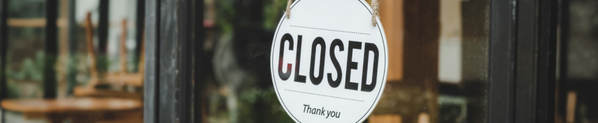 Nahaufnahme eines „Closed“-Schilds an einer Glastür, im Hintergrund ein geschlossenes Café oder Geschäft.