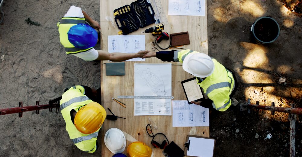 Overhead view of three construction workers wearing safety helmets, reviewing construction plans together at a table and coordinating on a building project.