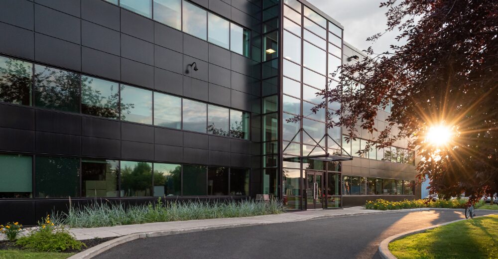 Modernes Bürogebäude mit Glasfassade im Abendlicht, vor dem Eingang Grünflächen und ein beleuchteter Zugangsbereich.