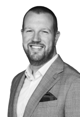 Black-and-white portrait of a smiling man with very short hair and a neatly trimmed beard. He is wearing a light-colored dress shirt, a textured blazer, and a pocket square. He is turned slightly to one side while looking directly at the camera. The background is plain and light-colored.