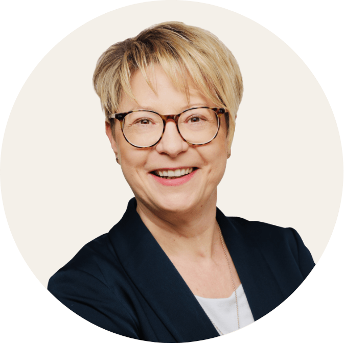 portrait of a smiling woman with short blond hair and round glasses. She is wearing a blazer and looking directly into the camera.