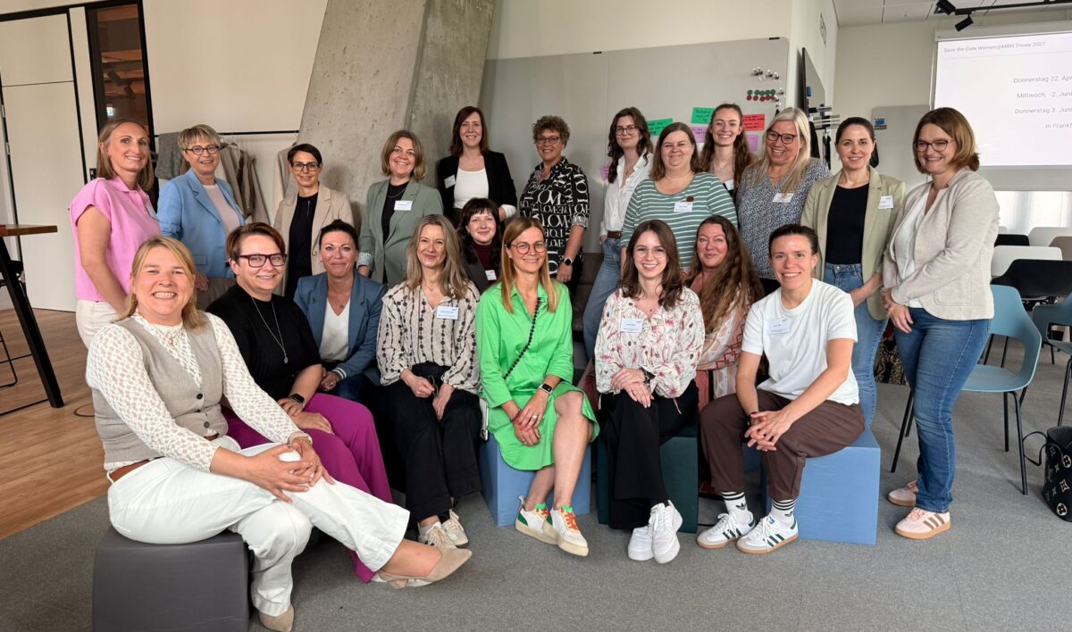 Gruppenfoto der Women@-Teilnehmerinnen bei einem gemeinsamen Treffen. Rund 20 Frauen unterschiedlichen Alters stehen und sitzen in mehreren Reihen zusammen und lächeln in die Kamera. Die Gruppe befindet sich in einem hellen, modernen Innenraum.