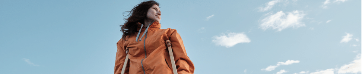 Eine Frau mit langen Haaren und einem orangefarbenen Jacke steht auf einem großen Felsen. Sie benutzt zwei Krücken zum Gehen. Der Hintergrund zeigt einen weiten Himmel mit einigen Wolken, die eine friedliche und offene Umgebung suggerieren.