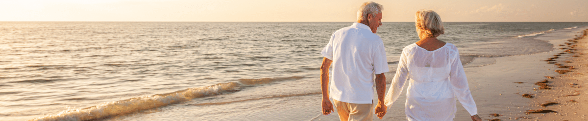 Älteres Paar in heller Kleidung geht Hand in Hand am Strand entlang. Die Sonne steht tief über dem Meer und taucht den Strand und das Wasser in warmes Licht.