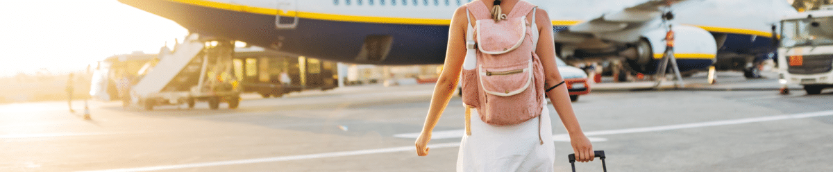Eine Frau mit Hut und Rucksack geht mit ihrem Gepäckkoffer über den Flughafen-Tarmac. Im Hintergrund steht ein großes Flugzeug bereit zum Abflug.