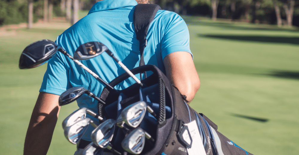 Ein Mann in einem blauen T-Shirt und einer schwarzen Kappe trägt einen Golfschläger-Rucksack auf einem Golfplatz.
