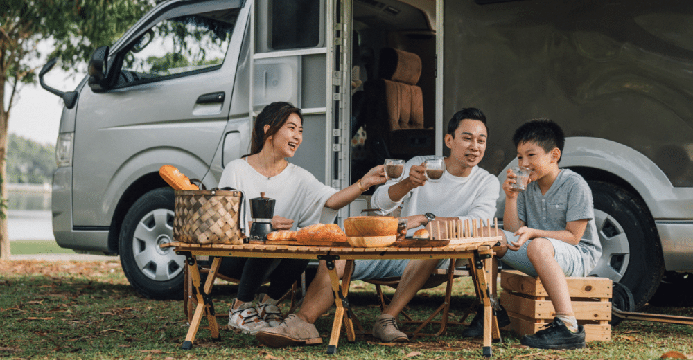 Eine Familie genießt ein Picknick vor ihrem Wohnmobil. Die Eltern und ihr Kind sitzen an einem Klapptisch und trinken aus Bechern, während sie Brot und andere Lebensmittel teilen.
