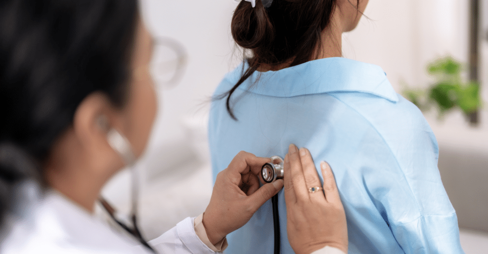 Eine Ärztin mit weißem Kittel und Stethoskop untersucht eine Patientin, die einen blauen Kittel trägt. Die Szene spielt sich in einem hellen, freundlichen Raum ab.