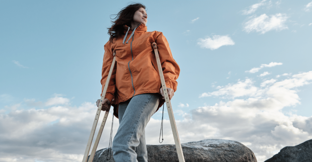 Eine Frau mit langen Haaren und einem orangefarbenen Jacke steht auf einem großen Felsen. Sie benutzt zwei Krücken zum Gehen. Der Hintergrund zeigt einen weiten Himmel mit einigen Wolken, die eine friedliche und offene Umgebung suggerieren.