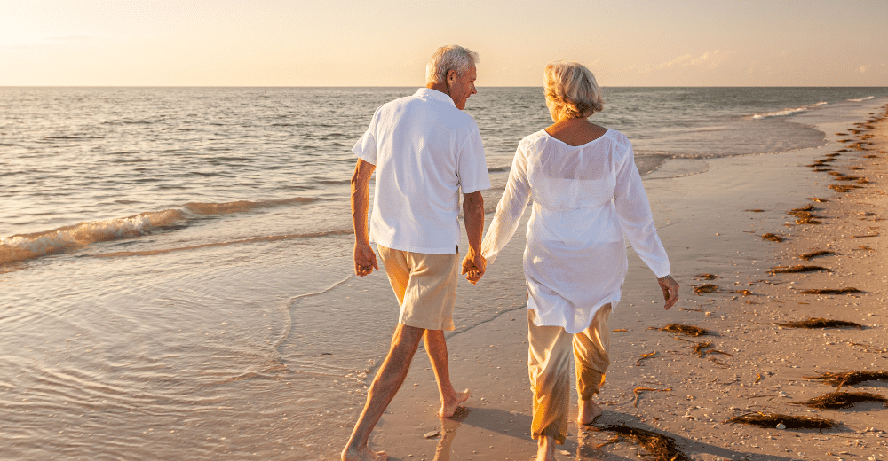 Älteres Paar in heller Kleidung geht Hand in Hand am Strand entlang. Die Sonne steht tief über dem Meer und taucht den Strand und das Wasser in warmes Licht.
