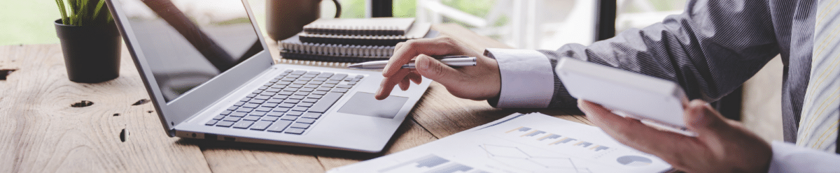Geschäftsperson in blauem Hemd arbeitet konzentriert an einem Holzschreibtisch mit geöffnetem Laptop und hält dabei einen Stift in der Hand. Auf der Arbeitsfläche liegen mehrere Notizbücher, Papiere mit Diagrammen und Grafiken, die auf geschäftliche Planung oder Datenanalyse deuten. Der moderne, helle Arbeitsplatz wird durch eine grüne Topfpflanze ergänzt und vermittelt eine produktive, professionelle Arbeitsumgebung mit natürlichem Tageslicht.