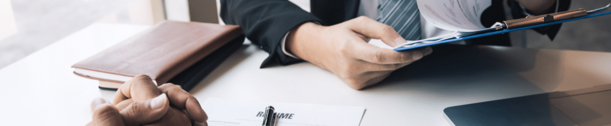 Two businesspeople are sitting across from each other at a white desk, intently studying documents during a professional meeting or job interview. The man in a black suit and gray tie is holding an application packet and a pen, while his counterpart listens attentively and takes notes.