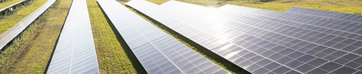 Aerial view of a solar farm in an open field: Dark photovoltaic modules are arranged in long, parallel rows, and the low-hanging sun is reflected on their surfaces. In the center stands a white battery storage container labeled “Energy Storage,” which complements the facility. The image illustrates a modern ground-mounted solar farm with energy storage and symbolizes renewable energy.