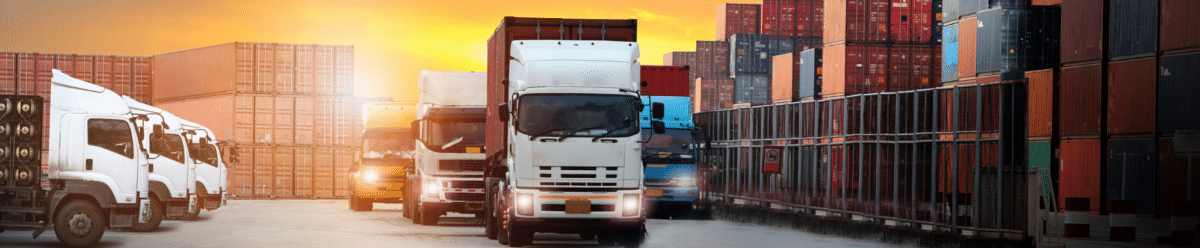 A logistics scene at sunset: In the foreground are several white and blue trucks in a concrete yard, one of them pulling a red container trailer. In the background, colorful containers (red, blue, gray, and orange) are stacked in tall rows, while two airplanes fly across the orange evening sky. The image symbolizes the connection between road, sea, and air freight.